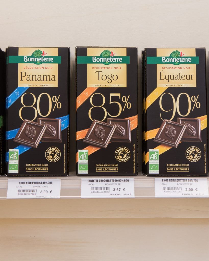 Tablettes de chocolat noir disposées sur un présentoir en bois avec porte-étiquettes transparents.